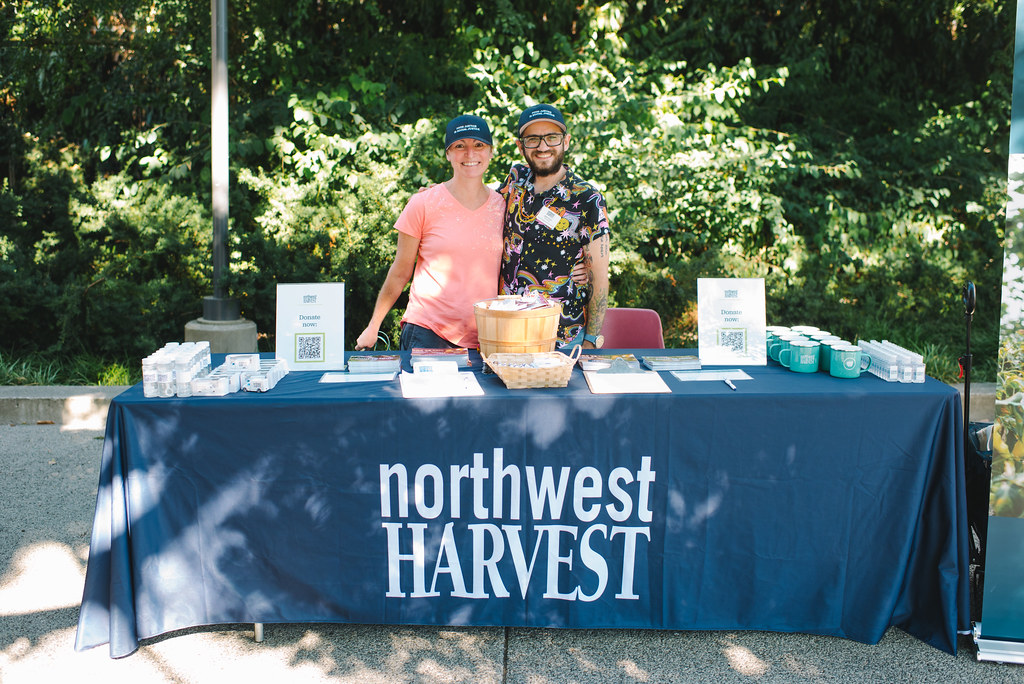 Two Northwest Harvest employees stand behind a table outside at a KEXP event