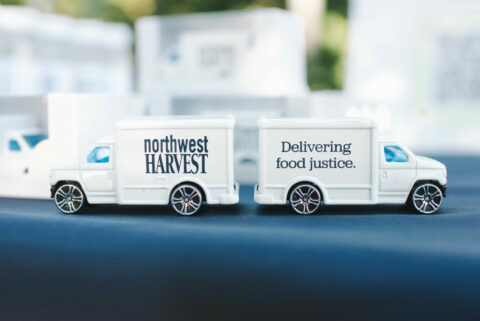 Photo of two toy box trucks facing end to end. Trucks are white with blue Northwest Harvest logo and message of 'delivering food justice'.