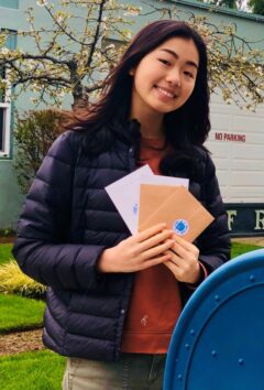 Hana from Hana Studios poses with envelopes held up in a fan shape while standing next to a USPS mailbox