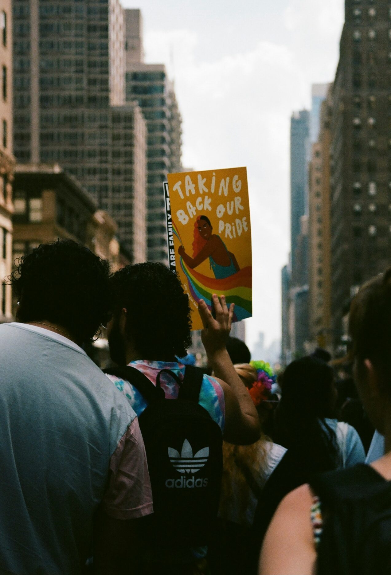 rear view of person holding up sign with text "Taking Back Our Pride" with graphic of a Black person grabbing a rainbow banner