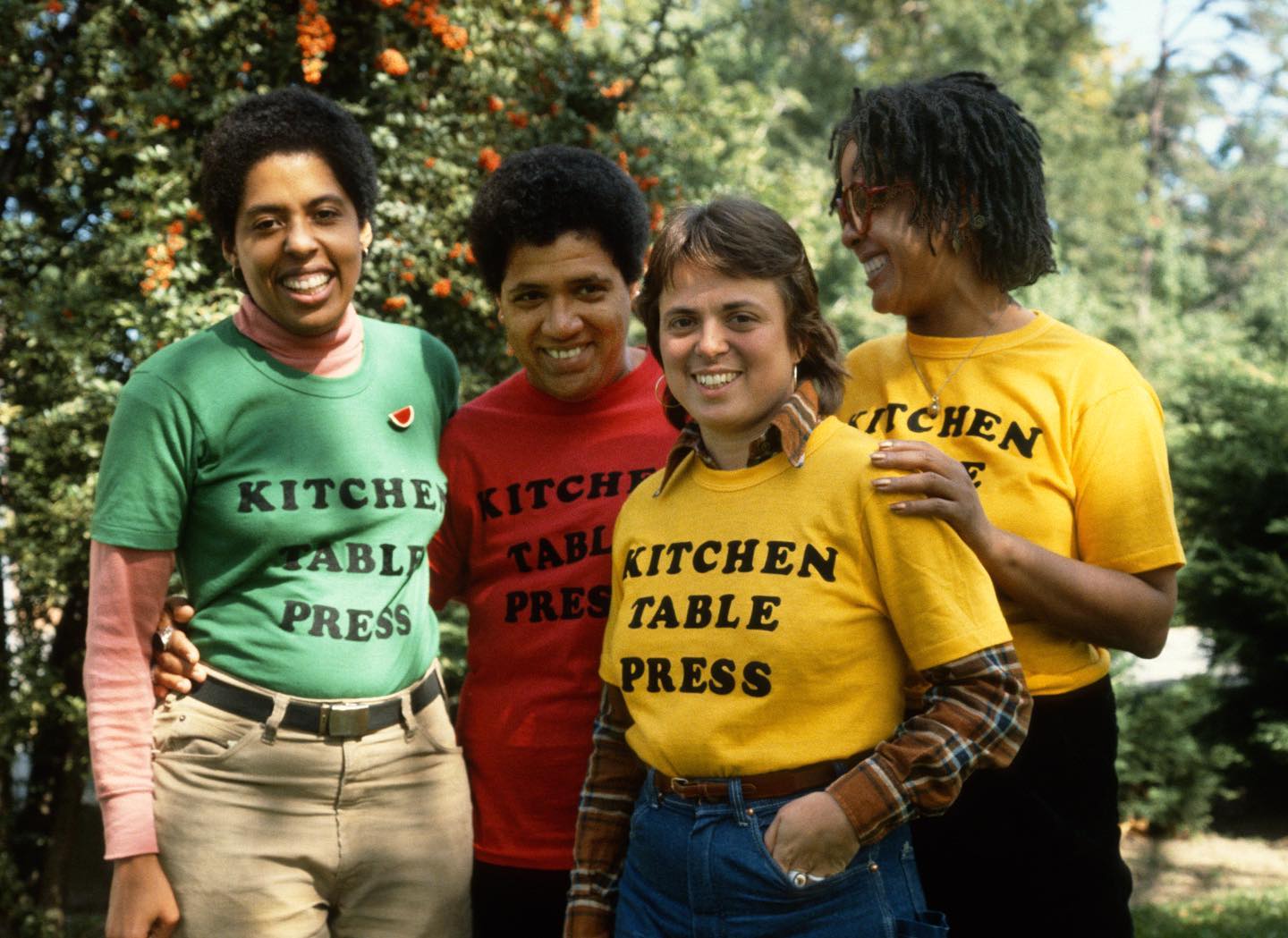 Group shot of members of Kitchen Table Press wearing green, red, and yellow shirts with the name of the press in black letters