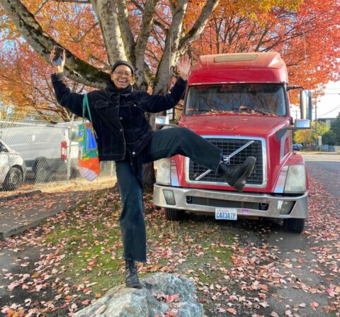 image of sol posing with arms up and left leg extended in front of red truck and tree with autumn leaves